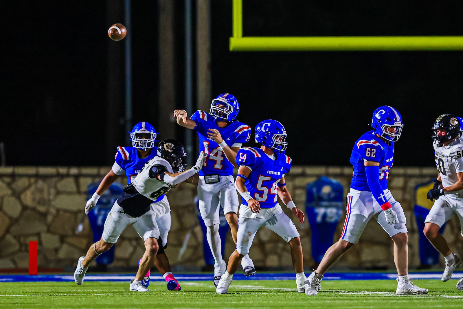 CT9A7939 | Press Pass Sports Blue-team football players on a night field; quarterback #14 throws a pass as teammates block and move downfield nearby defensemen.