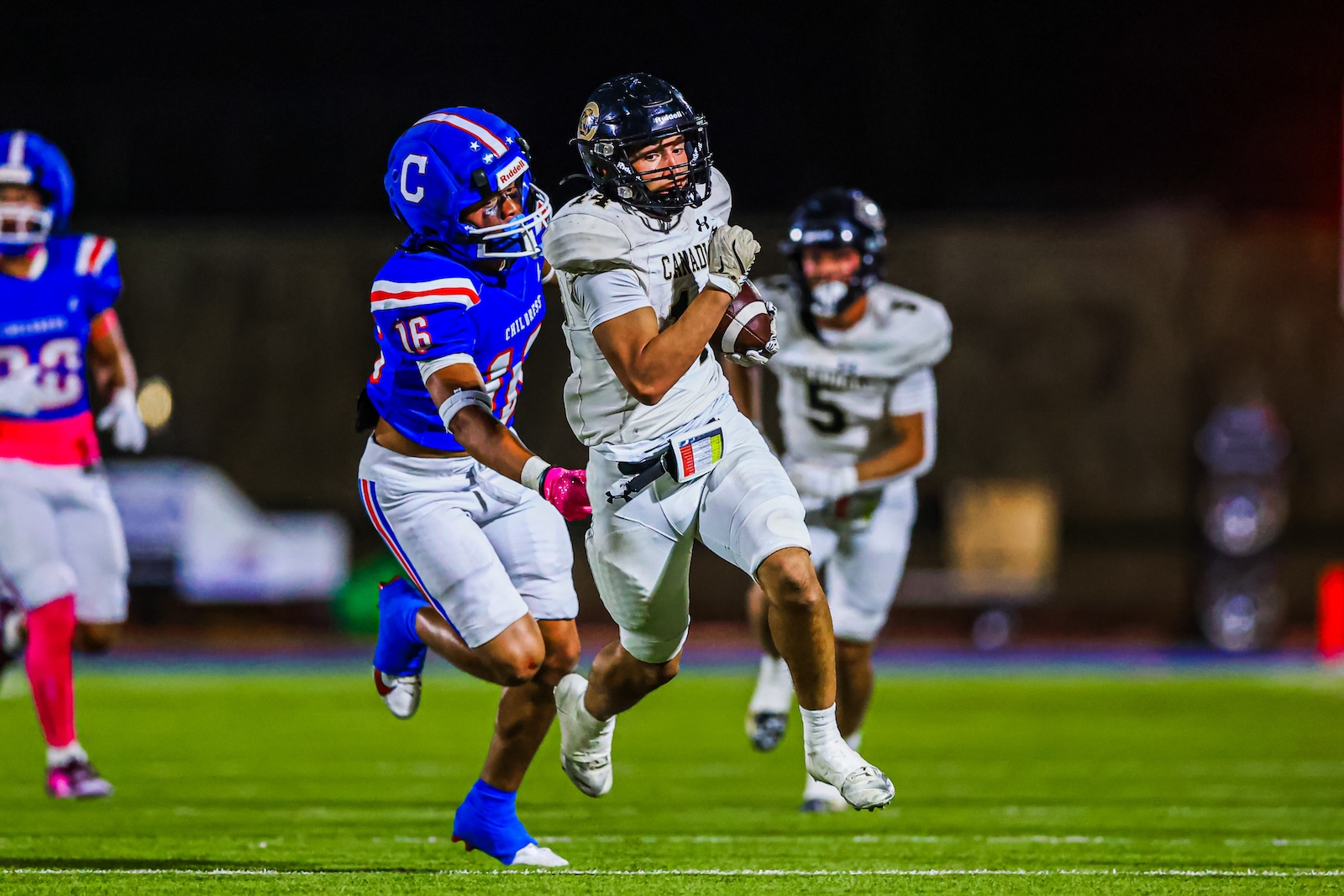 CT9A7977 | Press Pass Sports Running back in a white jersey carries the football while dodging a blue-clad defender at night on a turf field.