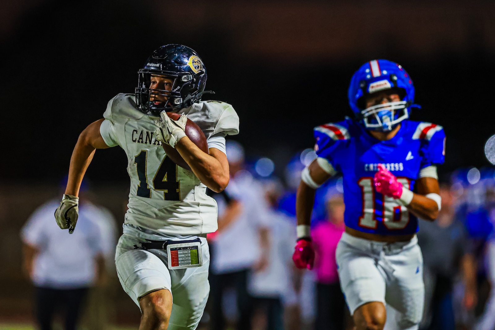 CT9A8059 | Press Pass Sports Two football players sprint on a night field; a white-jersey player wearing number 14 carries the ball while a blue-jersey defender number 18 pursues.