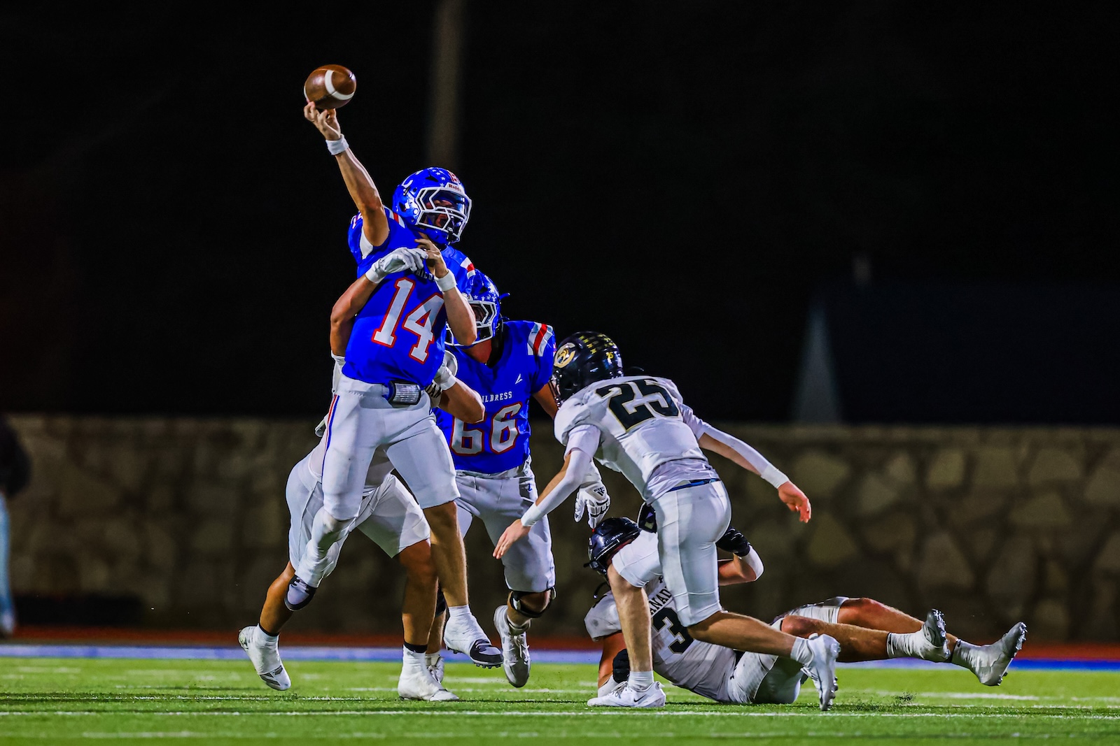 CT9A8149 | Press Pass Sports Football players in blue uniforms jump to catch a pass during a night game; defender in white tackles nearby on a green field.