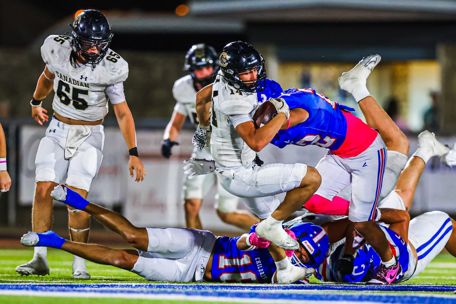 CT9A9246 | Press Pass Sports Tackle scene on a nighttime football field: players in white jerseys stop a blue jersey ball carrier holding the ball.