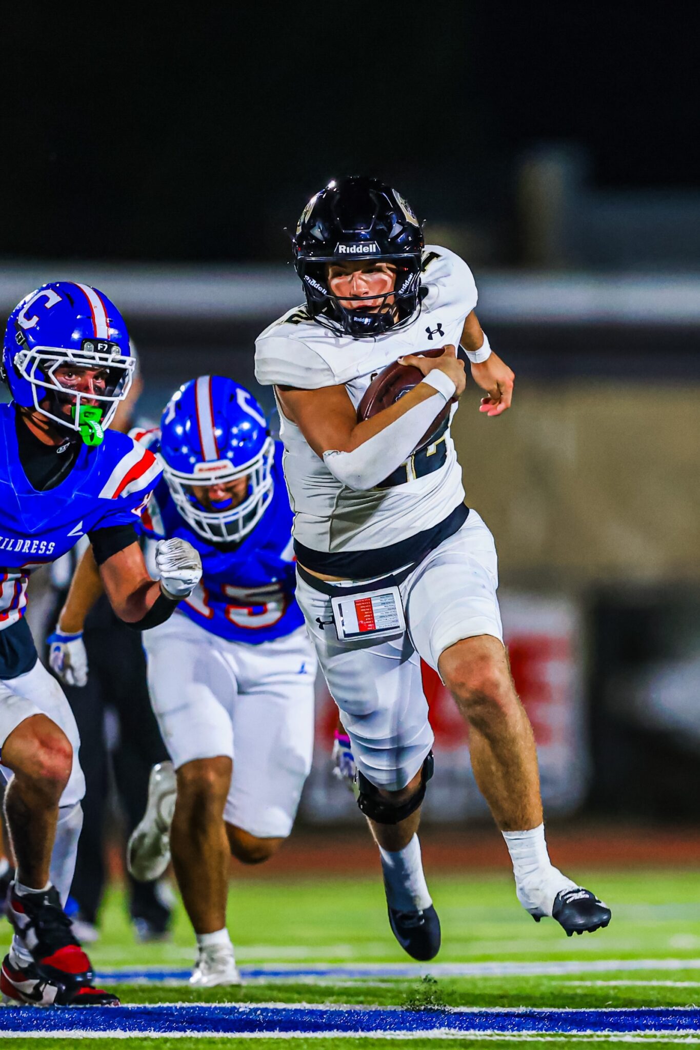 CT9A9282 | Press Pass Sports White-uniform football player carrying the ball runs forward while blue-clad defenders chase in the background on a night game field