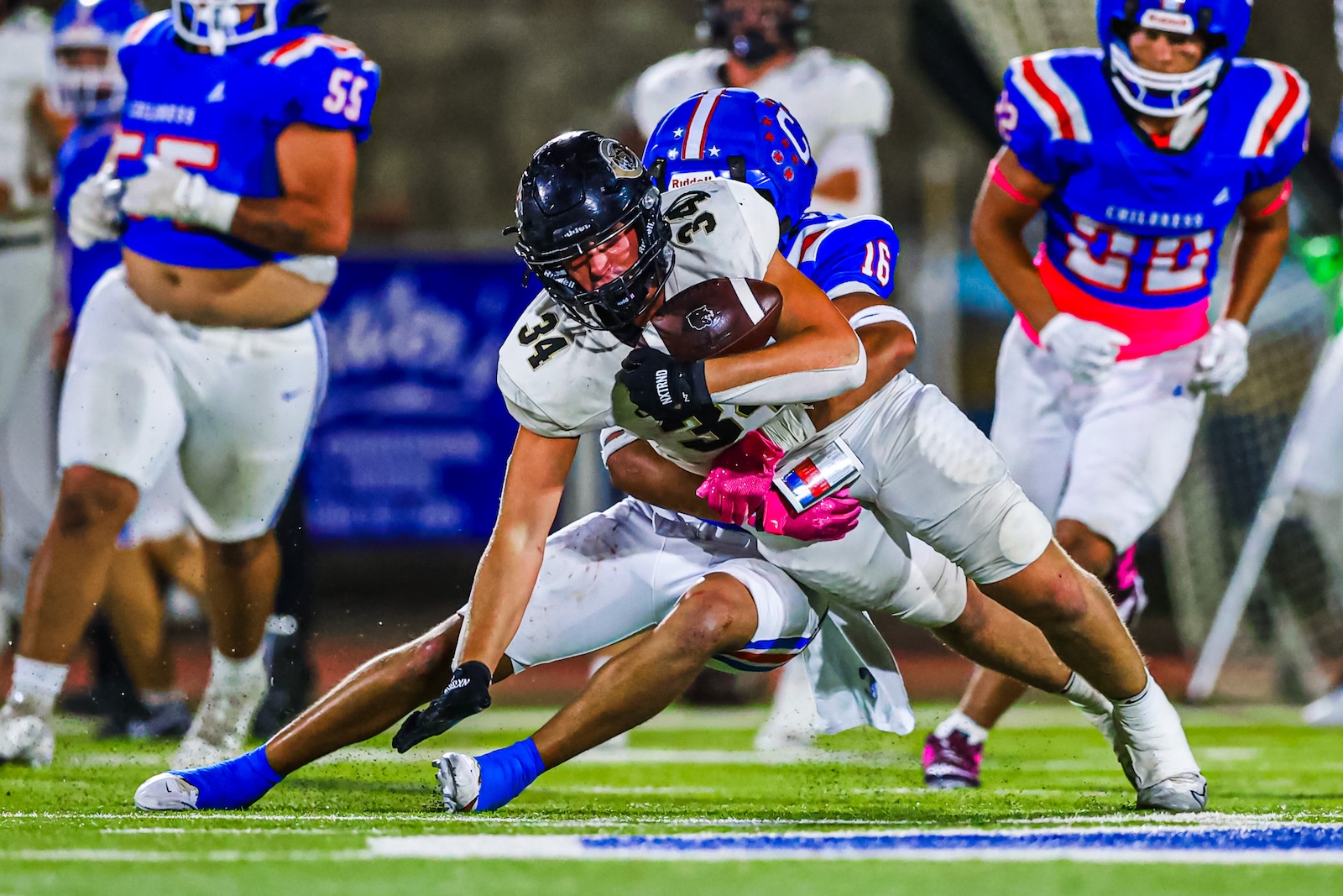 CT9A9542 | Press Pass Sports Football player in white uniform carrying the ball is tackled by a defender in blue, pink gloves visible on the tackler, on a bright turf field at night.