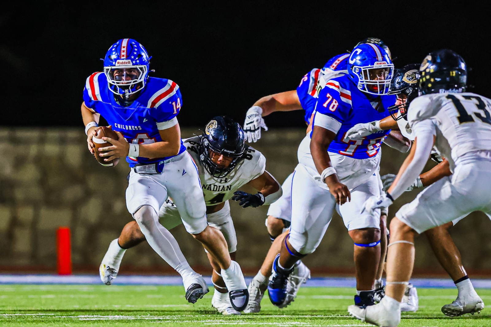 CT9A9776 | Press Pass Sports Blue team quarterback (#14) runs with the ball as white-clad defenders chase him on a night game turf scene.