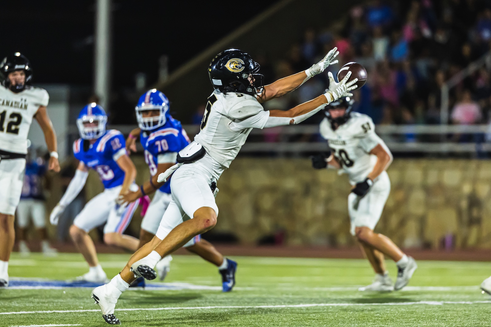 DE3_1174 | Press Pass Sports Football player in white reaching for a caught ball during a night game, with teammates in blue in the background and a crowd watching.