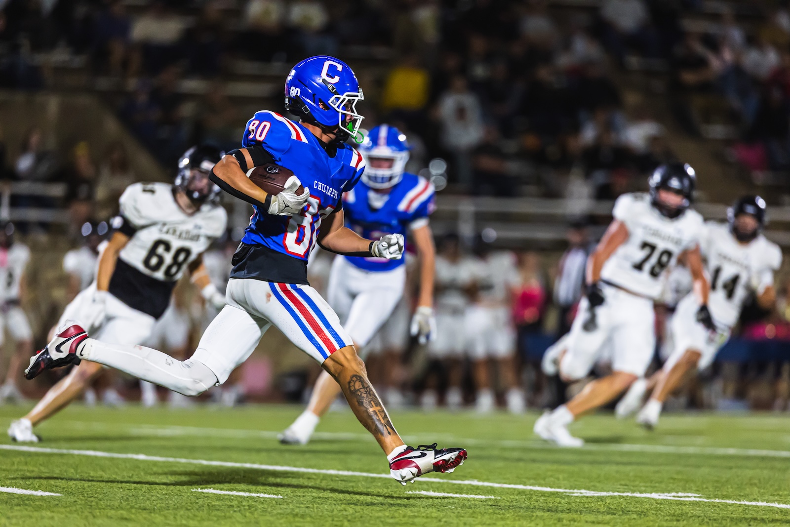DE3_1747 | Press Pass Sports Blue-team running back with a'C' captain helmet carries the ball while sprinting past white-team defenders on a night game field.