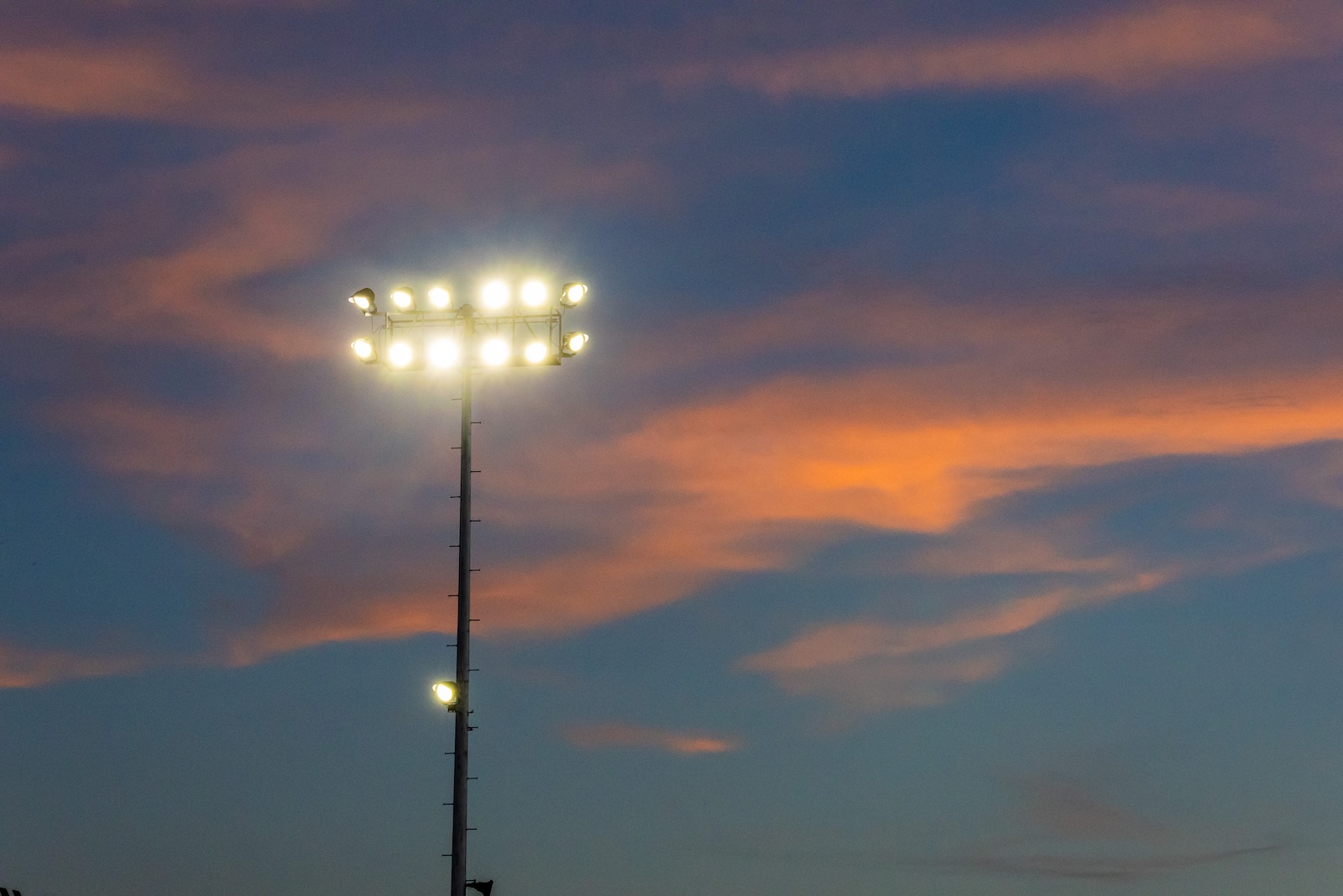 DE3_8311 | Press Pass Sports Tall floodlight tower with bright stadium lights against a sunset sky at dusk.