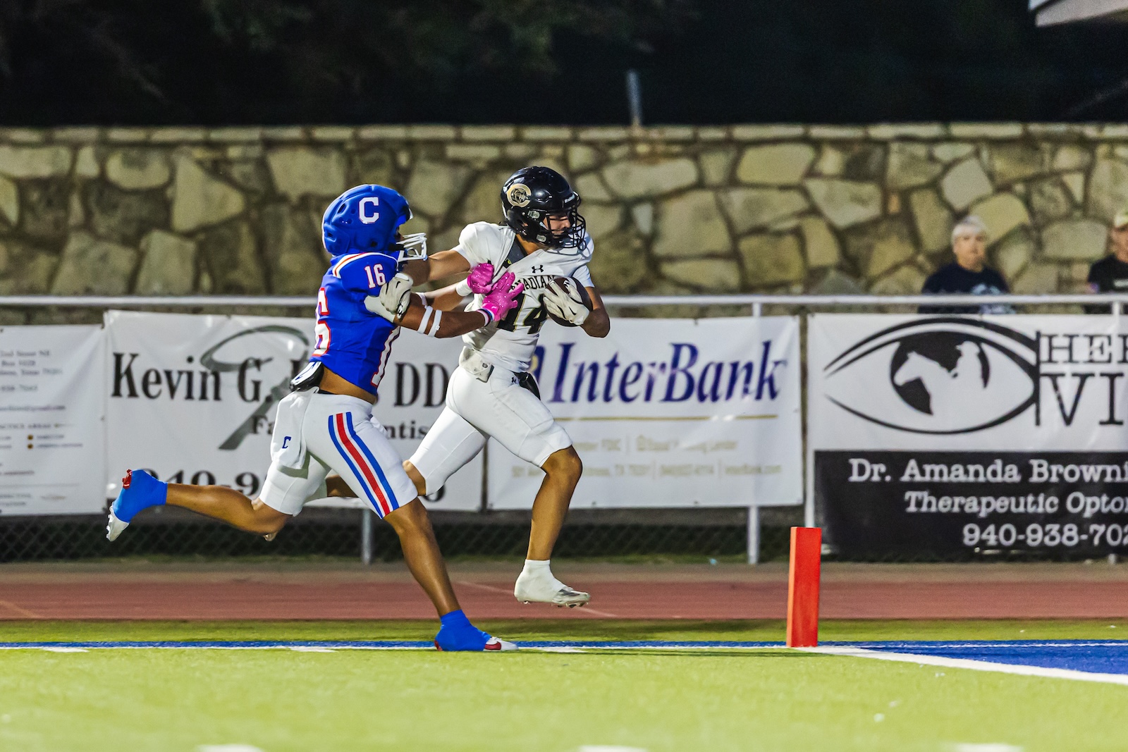 DE3_8848 | Press Pass Sports Two football players in a night game: a white-uniform receiver leaping with the ball as a blue-uniform defender closes in.