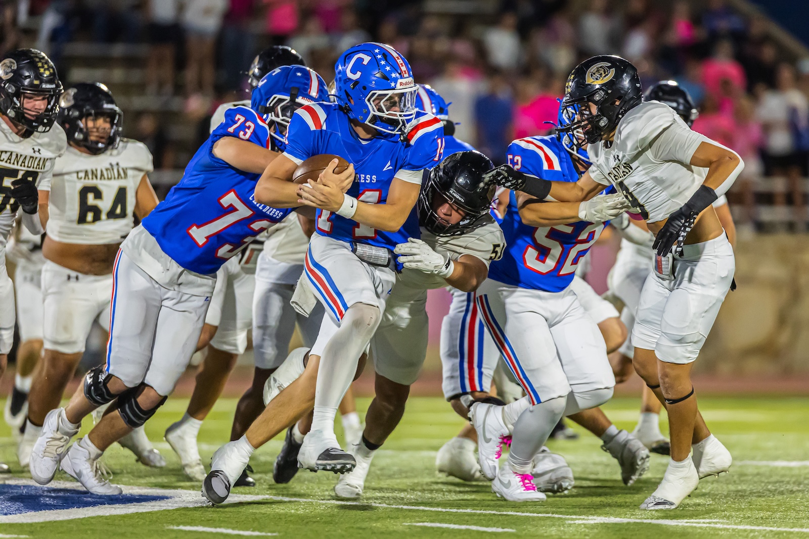 DE3_9063 | Press Pass Sports Blue-uniform quarterback cradles the ball as defenders in white close in during a tense football play at night.