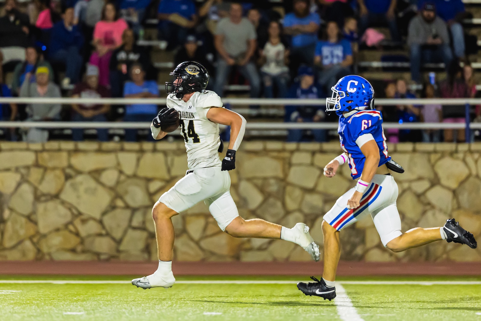 DE3_9374 | Press Pass Sports Football player in white jersey runs with ball on a green field, chased by a defender in blue jersey amid a crowded stadium.