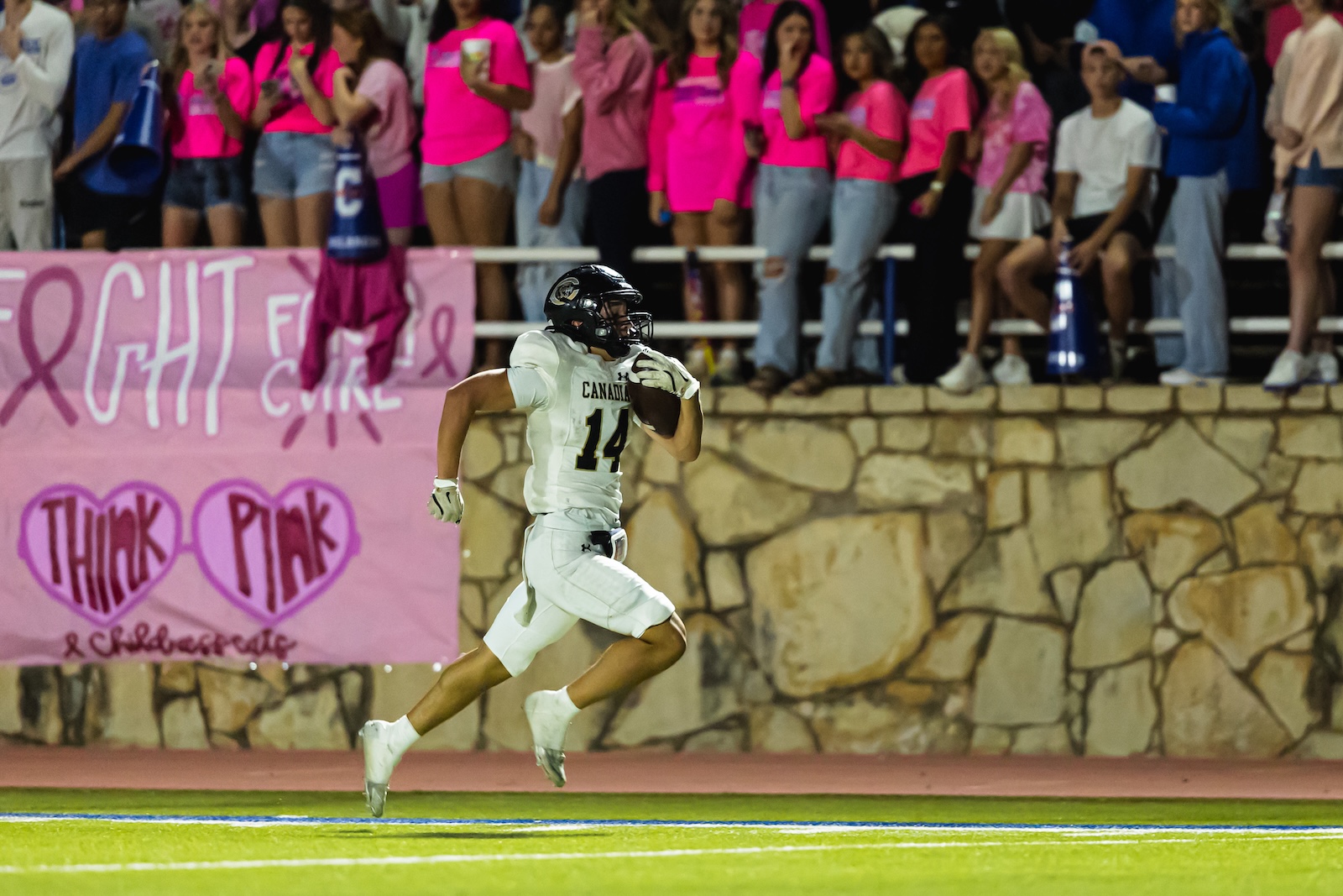DE3_9753 | Press Pass Sports Canadian football player wearing number 14 in white runs with the ball on a green field, crowd in pink shirts cheering in the stands behind a pink breast cancer banner.