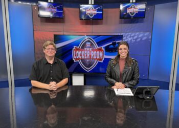 Two news anchors sit at a glossy desk in a blue TV studio with screens showing The Locker Room logo behind them.