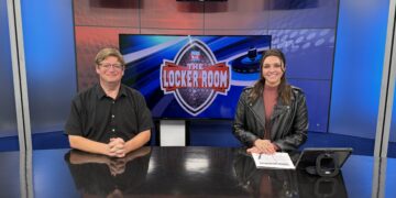 Two news anchors sit at a glossy desk in a blue TV studio with screens showing The Locker Room logo behind them.