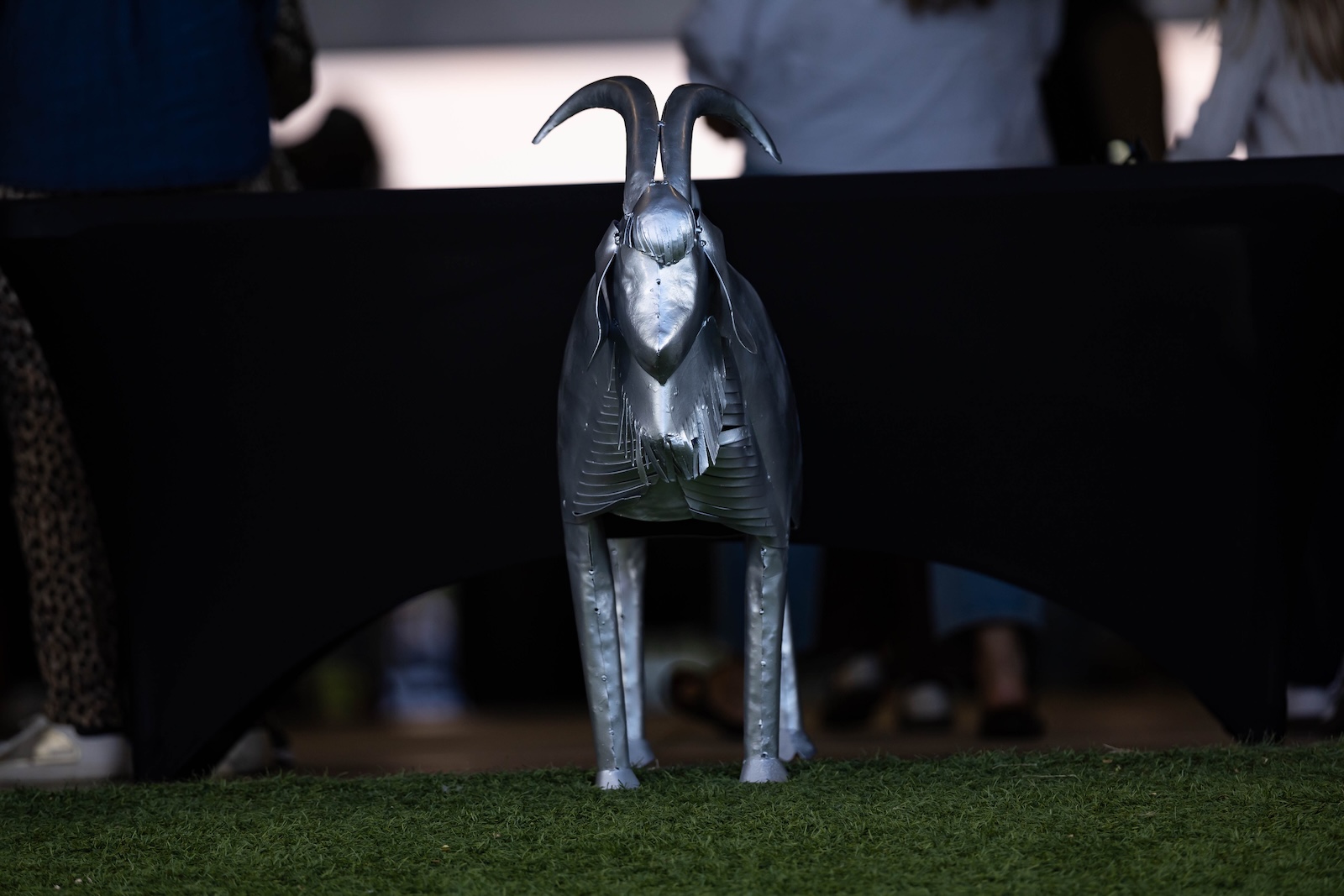 RHS-CHS-FB-1 | Press Pass Sports Metallic goat sculpture standing on grass in front of a dark table, horns curved upward, futuristic metallic design.