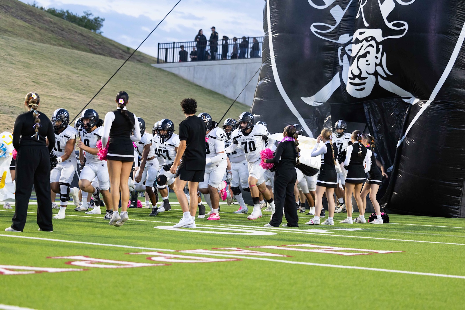 RHS-CHS-FB-3 | Press Pass Sports Football players in white uniforms rush onto the field from a large black inflatable tunnel with a Raider mascot, cheerleaders nearby on turf.