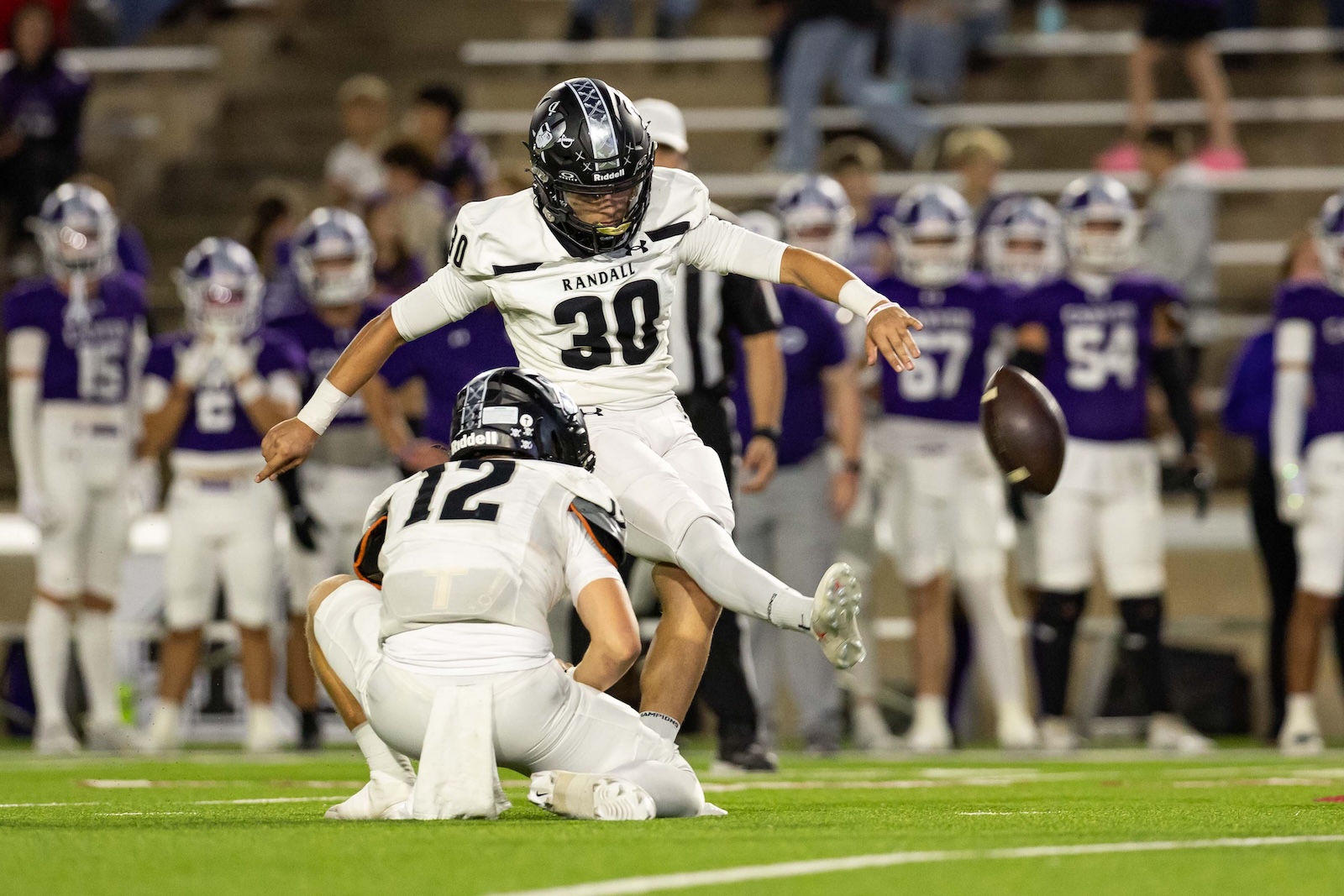 RHS-CHS-FB-12 | Press Pass Sports White-uniform football player #30 leaps over a tackle as a football flies nearby on a green field; purple-clad players sit on the sideline in the background.