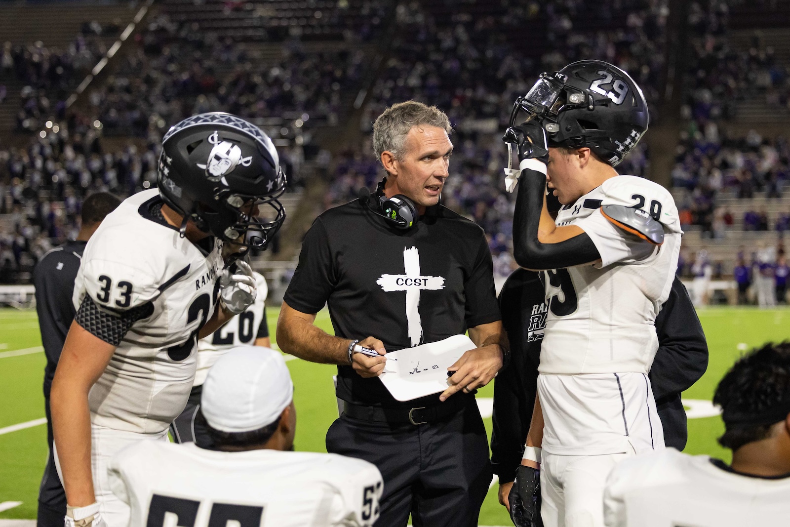 RHS-CHS-FB-13 | Press Pass Sports Coach on the sideline talks to two players in white uniforms during a football game, headset around his neck.