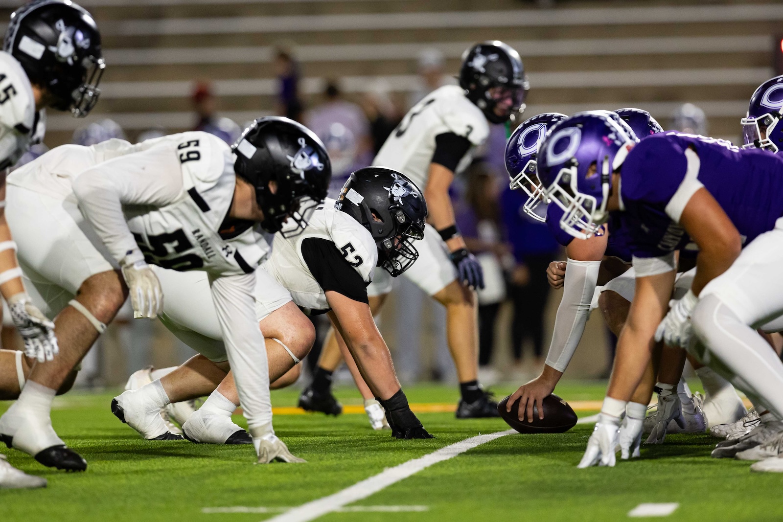 RHS-CHS-FB-14 | Press Pass Sports Line of scrimmage in a high school football game, center crouched to snap the ball between white and purple teams on a green turf field.