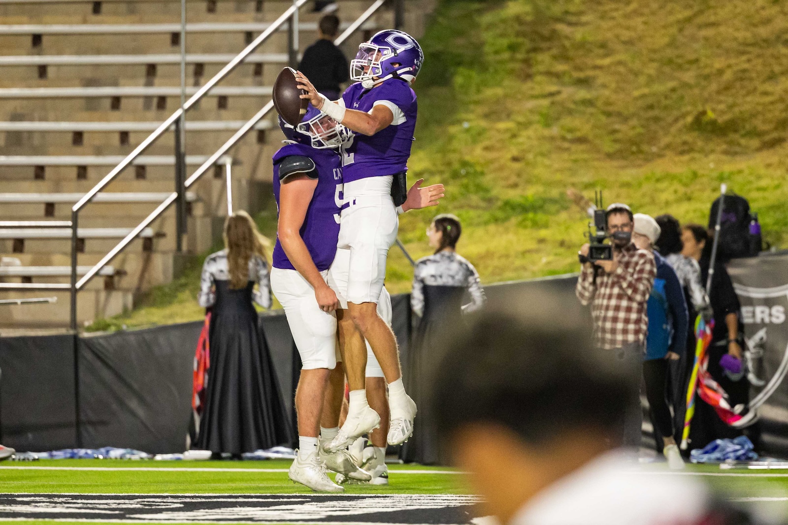 RHS-CHS-FB-16 | Press Pass Sports Two football players in purple and white uniforms celebrate on the field as one jumps to catch the ball while a teammate supports him nearby. Behind them, photographers and a crowd watch near the sidelines.