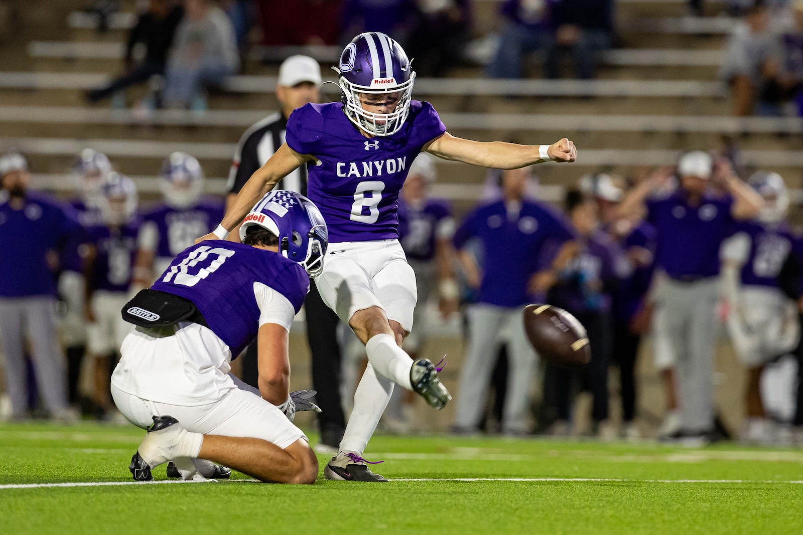 RHS-CHS-FB-18 | Press Pass Sports Purple-clad football player (no. 8) leaps to kick the ball as a teammate blocks and a defender falls on the field behind him.