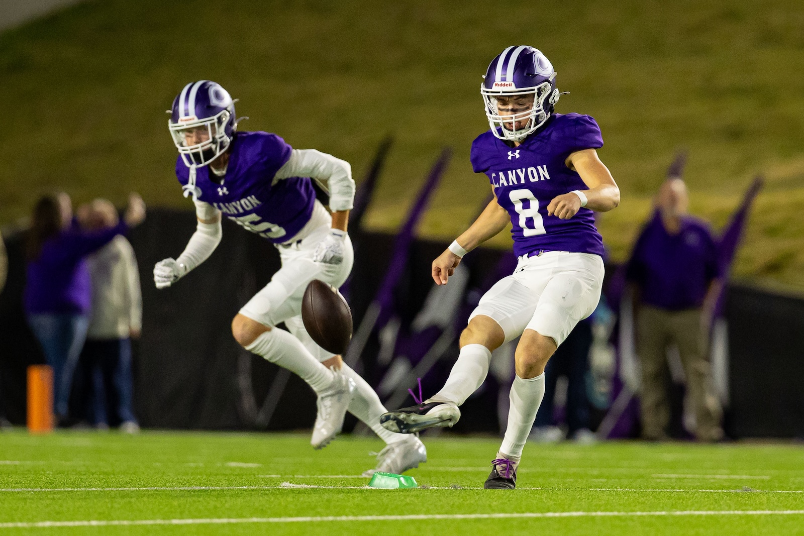 RHS-CHS-FB-19 | Press Pass Sports Two Canyon football players in purple uniforms on a night game; one kicks the ball while a teammate watches nearby.