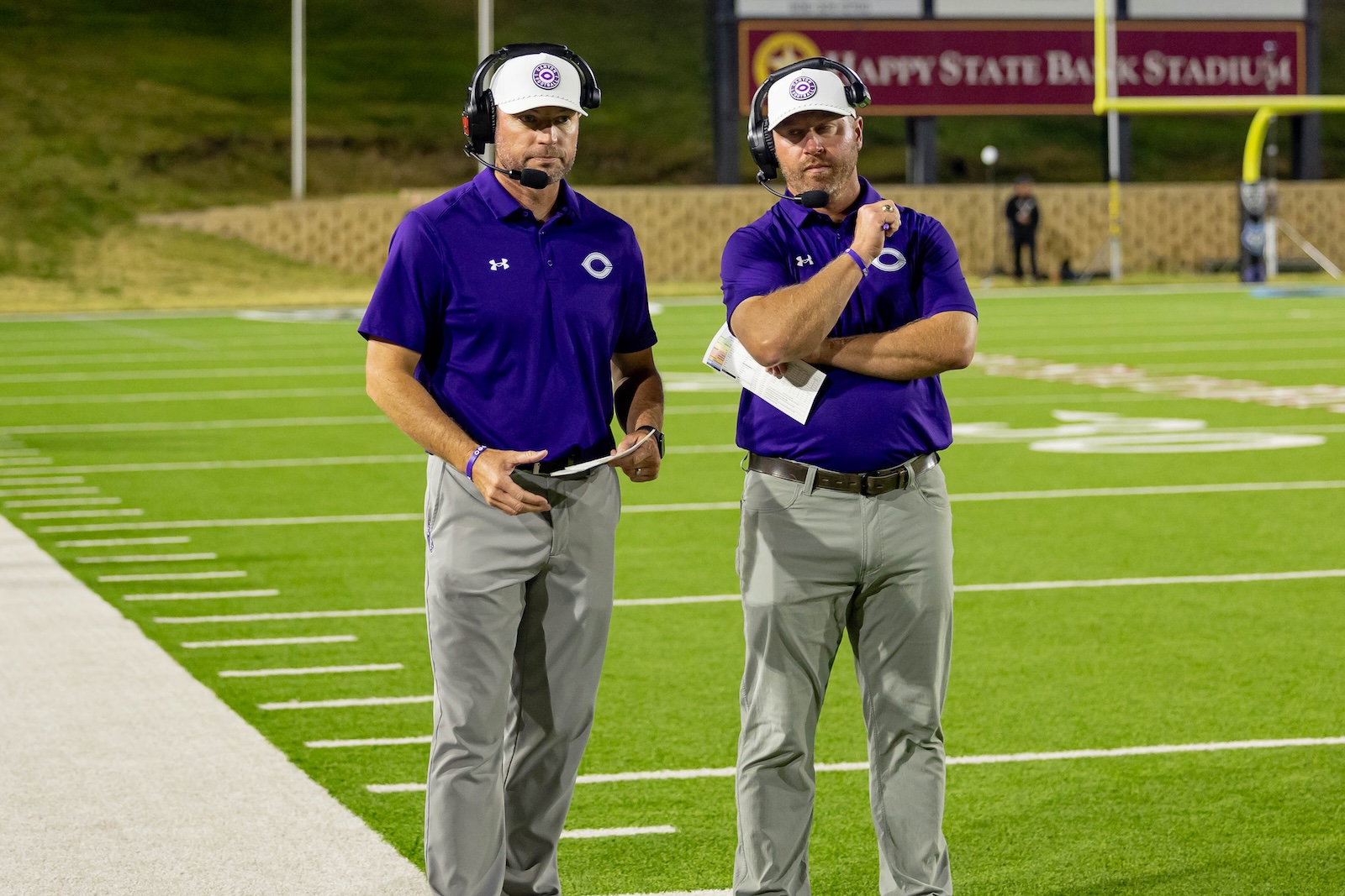 RHS-CHS-FB-23 | Press Pass Sports Two men in purple polo shirts with headsets stand on a football field, holding papers and looking toward the camera.