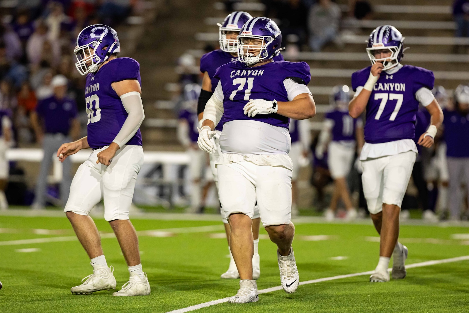 RHS-CHS-FB-24 | Press Pass Sports Three Canyon football players in purple jerseys walk on the green field during a game, numbers 50, 71, and 77 visible.