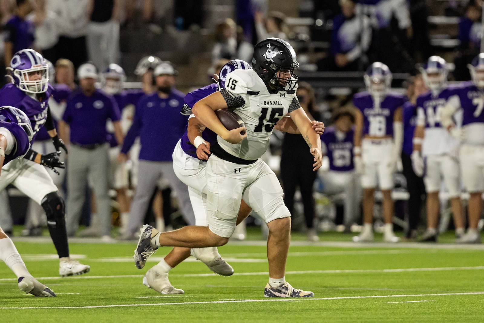 RHS-CHS-FB-26 | Press Pass Sports Football player wearing white, #15, runs with the ball as a purple-clad defender pursues on a green field at dusk.
