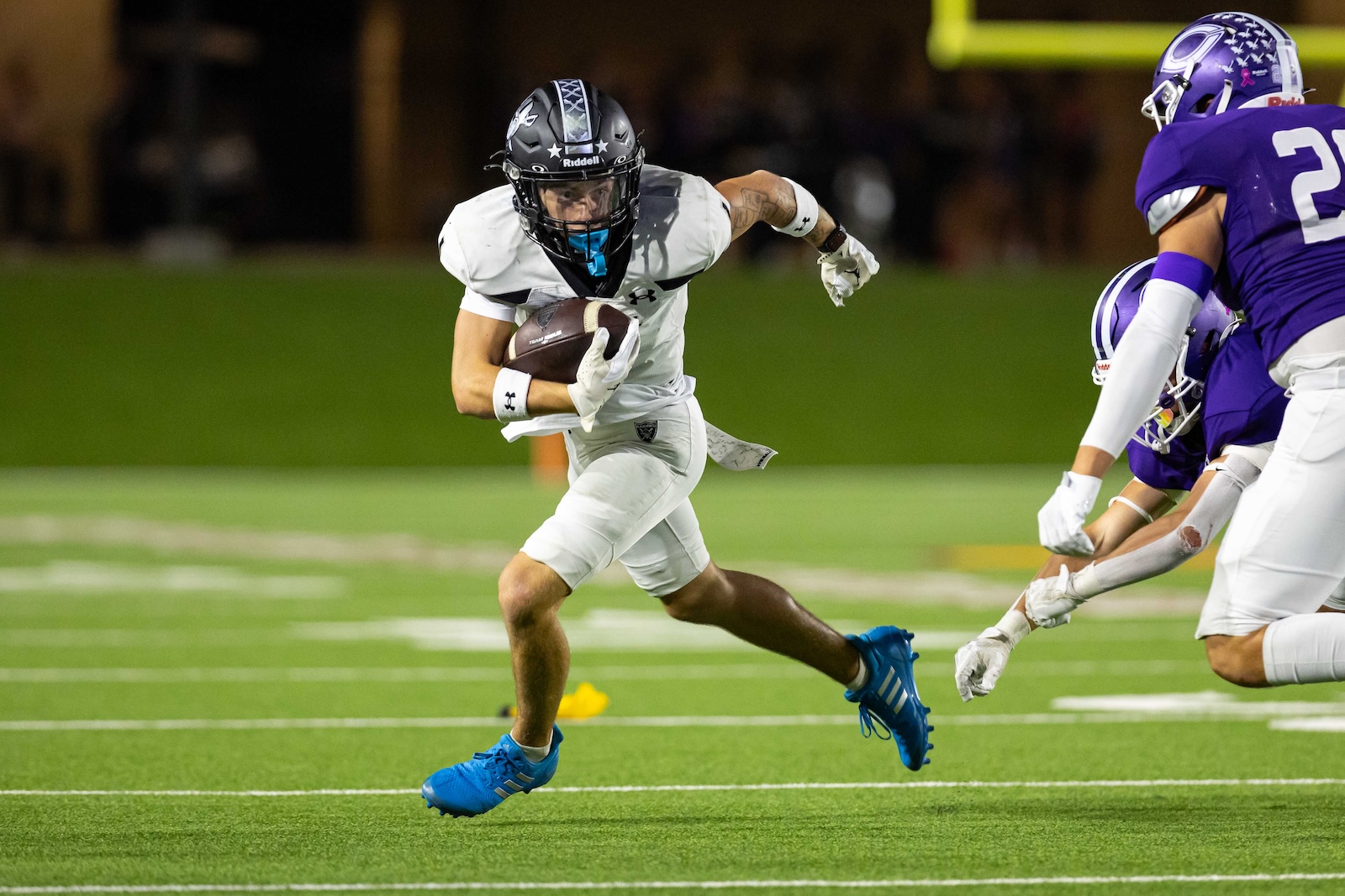 RHS-CHS-FB-27 | Press Pass Sports Football player in white jersey running with the ball while defenders in purple close in on a green field at night stadium pale background behind them.