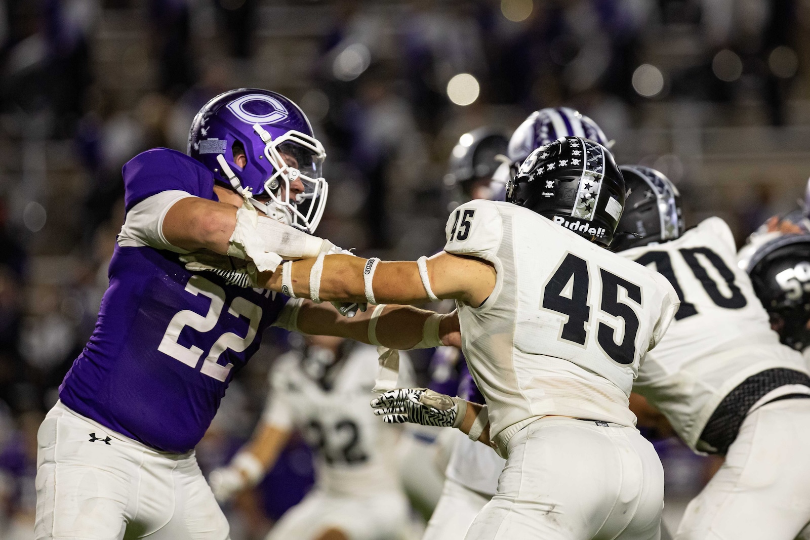 RHS-CHS-FB-32 | Press Pass Sports Two football players collide as white #45 blocks a purple defender (#25) during a game, crowd blurred in the background, night stadium lighting.