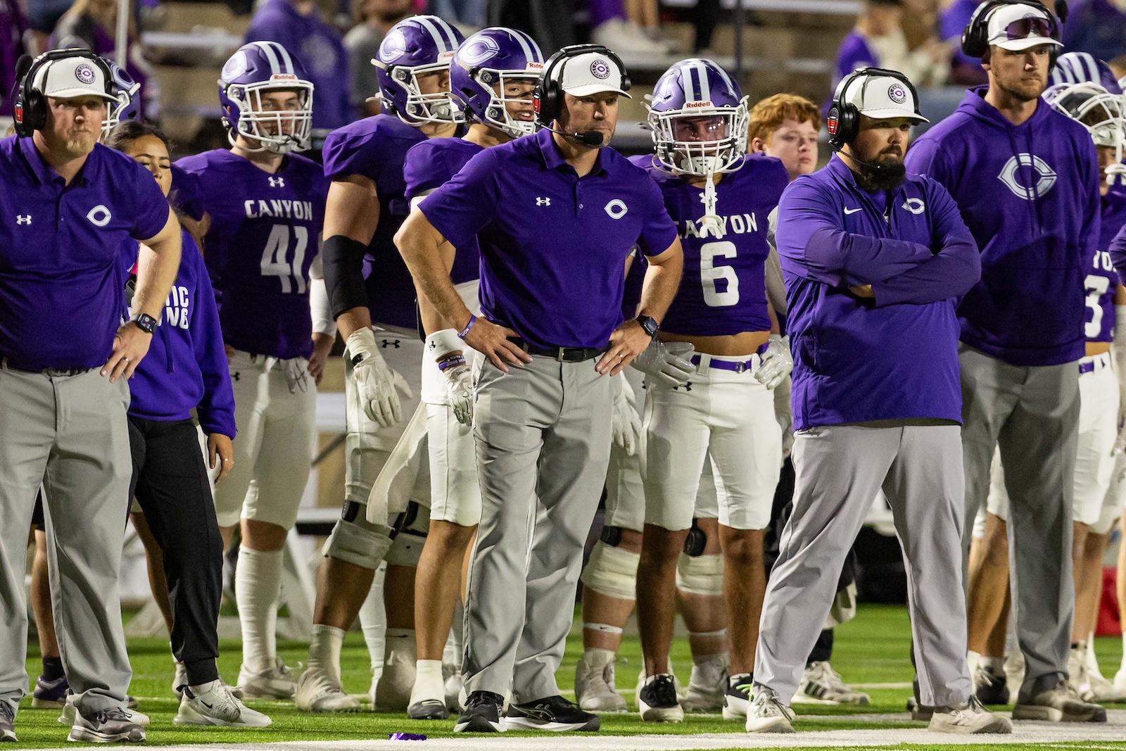 RHS-CHS-FB-36 | Press Pass Sports Canyon football coaches and players in purple uniforms stand with arms crossed on the sideline, helmets on and off, on a green field with a crowd in the background.