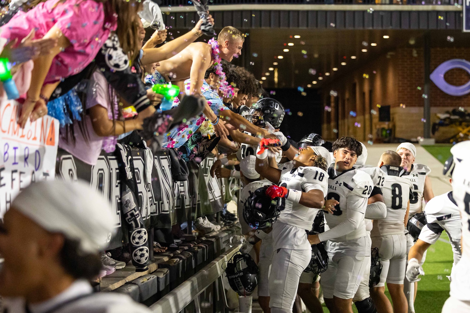 RHS-CHS-FB-38 | Press Pass Sports Football players in white uniforms high-five cheering fans from the sideline during a celebration, confetti in the air.