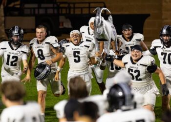 Randall football team celebrates victory on field, lifting a silver horse trophy together.