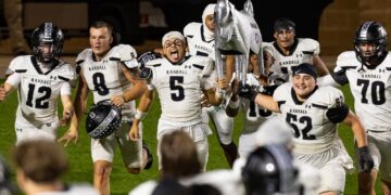 Randall football team celebrates victory on field, lifting a silver horse trophy together.