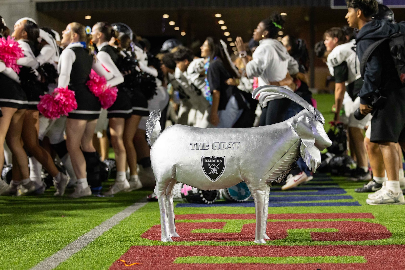 RHS-CHS-FB-40 | Press Pass Sports Silver inflatable goat with 'THE GOAT' and Raiders logo stands on a football field as a crowd of cheerleaders and players line up in the background.