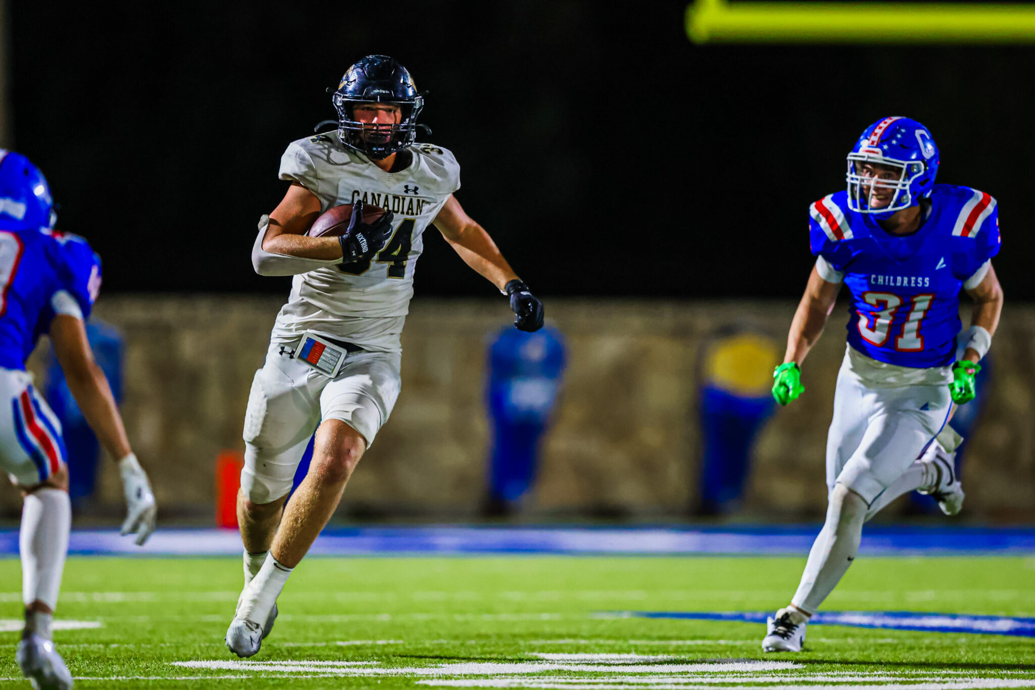 CT9A4515 | Press Pass Sports A football player in a white Canadian jersey runs with the ball on a night game while defenders in blue close in behind him.