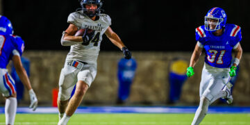 A football player in a white Canadian jersey runs with the ball on a night game while defenders in blue close in behind him.