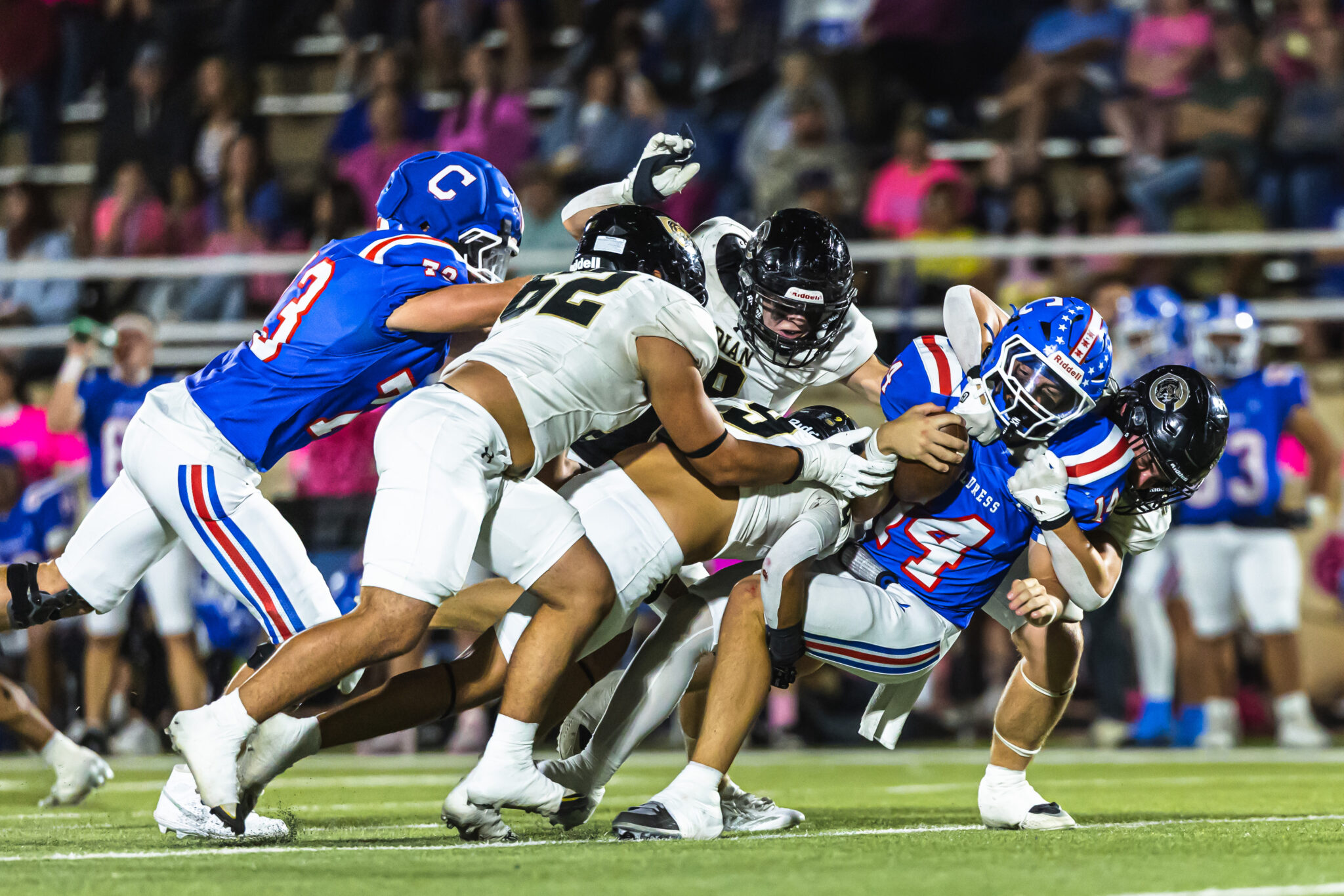 Players in blue and white uniforms clash as they dive to the ground during a football tackle on a crowded field. [action-packed moment]