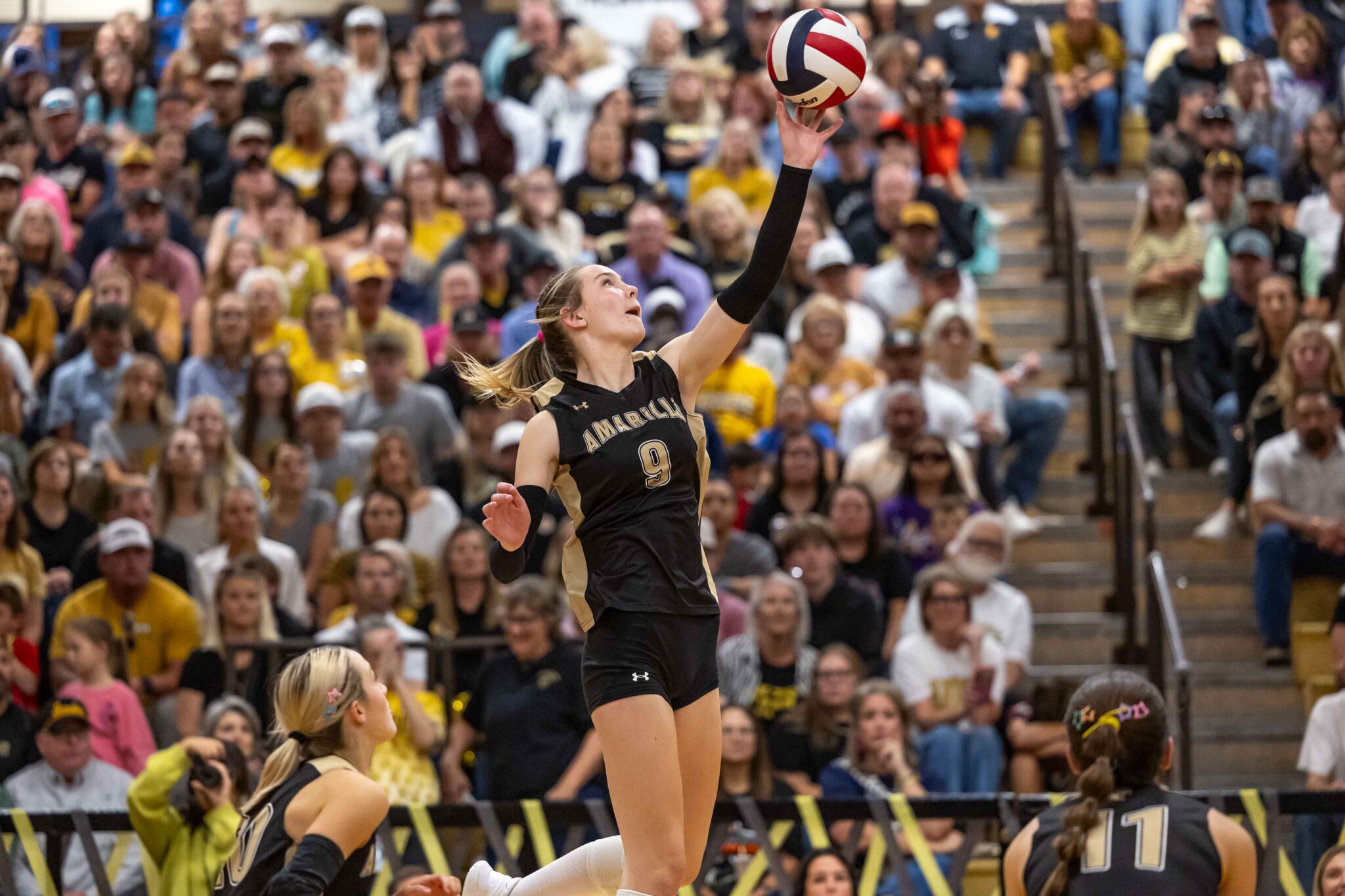 THS-AHS-VB-31 1 | Press Pass Sports Volleyball player in a black uniform with the number 9 jumps to spike the ball amid a cheering crowd in a gymnasium.