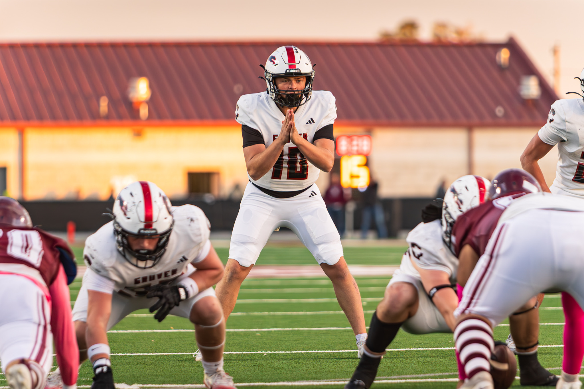 American football players in white uniforms form a huddle at the line of scrimmage on a green turf field during a sunset game, central player with hands pressed together in prayer-like gesture ready to snap or call a play.