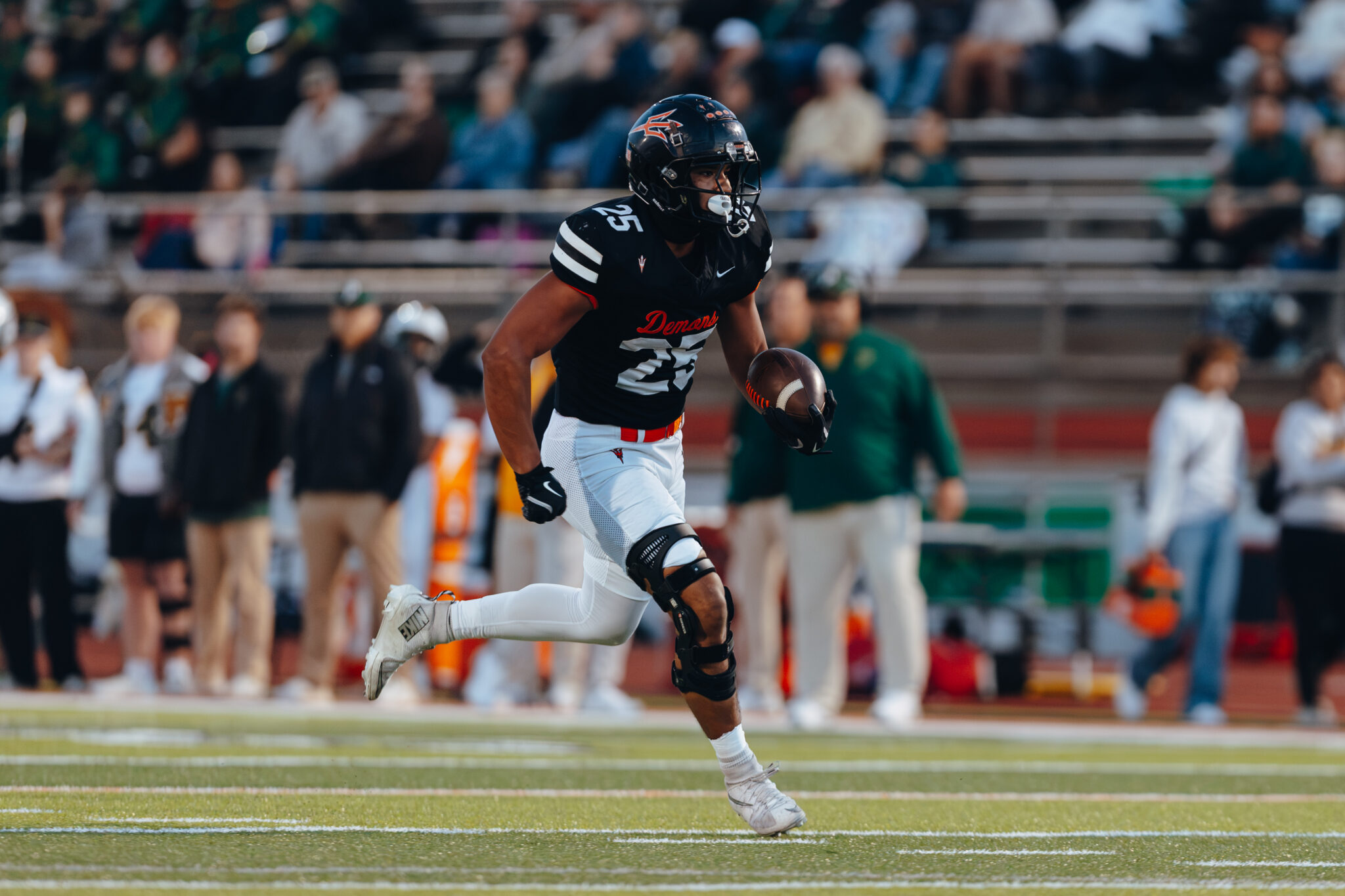 2C5A8014 | Press Pass Sports Football player in a black jersey (number 25) runs with the ball during a game, with a blurred crowd in the stands behind him, on a grass field.