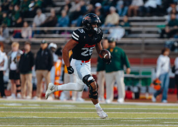 2C5A8014 | Press Pass Sports Football player in a black jersey (number 25) runs with the ball during a game, with a blurred crowd in the stands behind him, on a grass field.