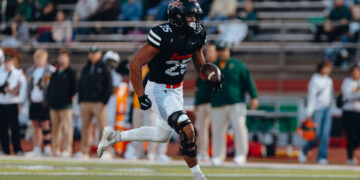 Football player in a black jersey (number 25) runs with the ball during a game, with a blurred crowd in the stands behind him, on a grass field.