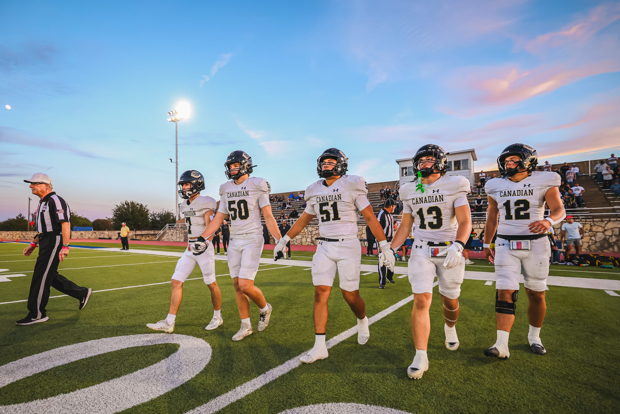 Canadian football players in white uniforms walk off the field together as a referee looks on, with bleachers and a sunset sky in the background