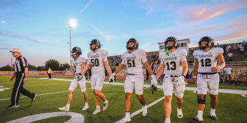 Canadian football players in white uniforms walk off the field together as a referee looks on, with bleachers and a sunset sky in the background