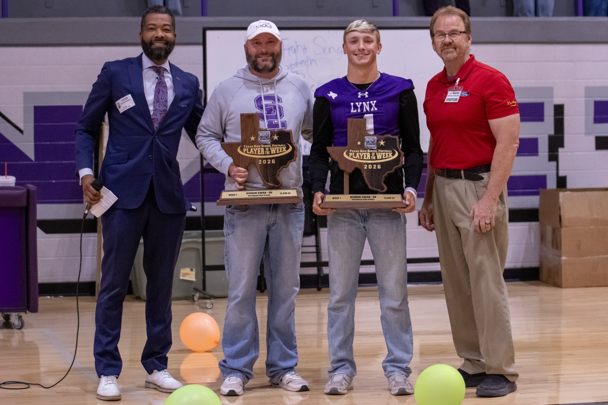 Four men stand on a gym stage posing with Texas-shaped 'Player of the Week 2026' trophies; balloons on the floor suggest a celebration.