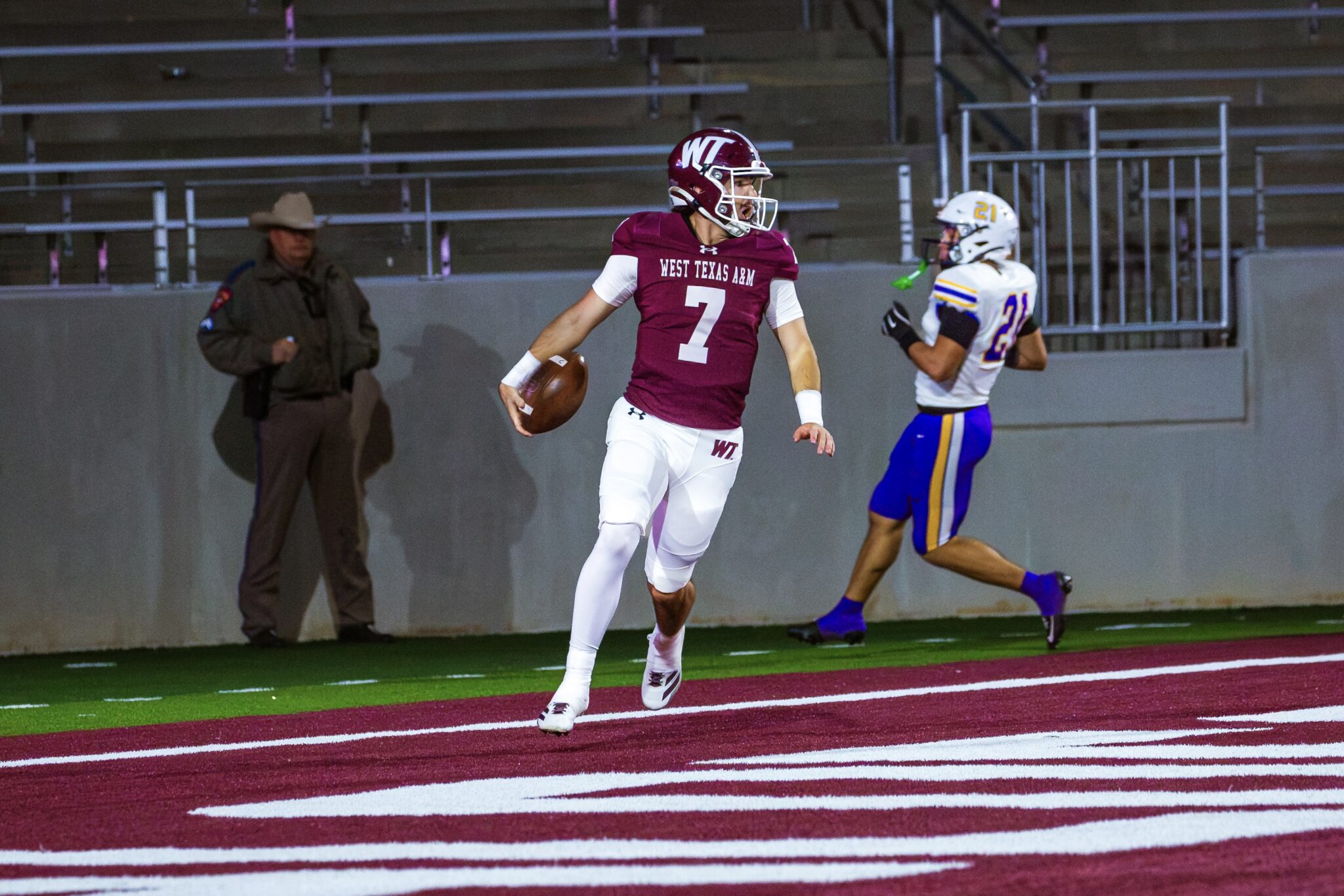 _V4A3296 | Press Pass Sports Quarterback in maroon West Texas A&M jersey #7 holding a football while running on a red turf field, pursued by a defender in white and blue.