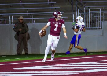 _V4A3296 | Press Pass Sports Quarterback in maroon West Texas A&M jersey #7 holding a football while running on a red turf field, pursued by a defender in white and blue.