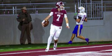 Quarterback in maroon West Texas A&M jersey #7 holding a football while running on a red turf field, pursued by a defender in white and blue.