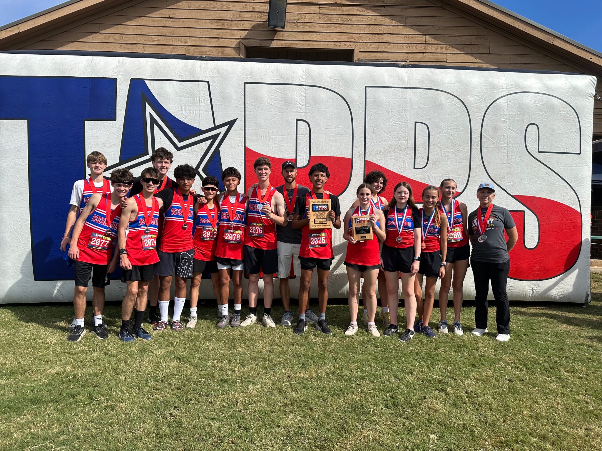 image | Press Pass Sports Youth track team posing with medals and trophies in front of a large banner on a sunny day.